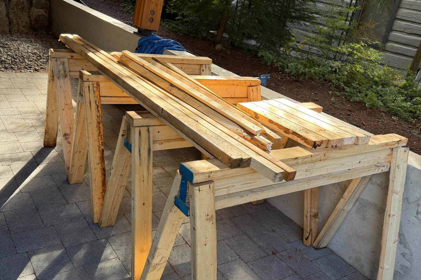 Two wooden sawhorses on a patio, stacked with cut lengths of lumber