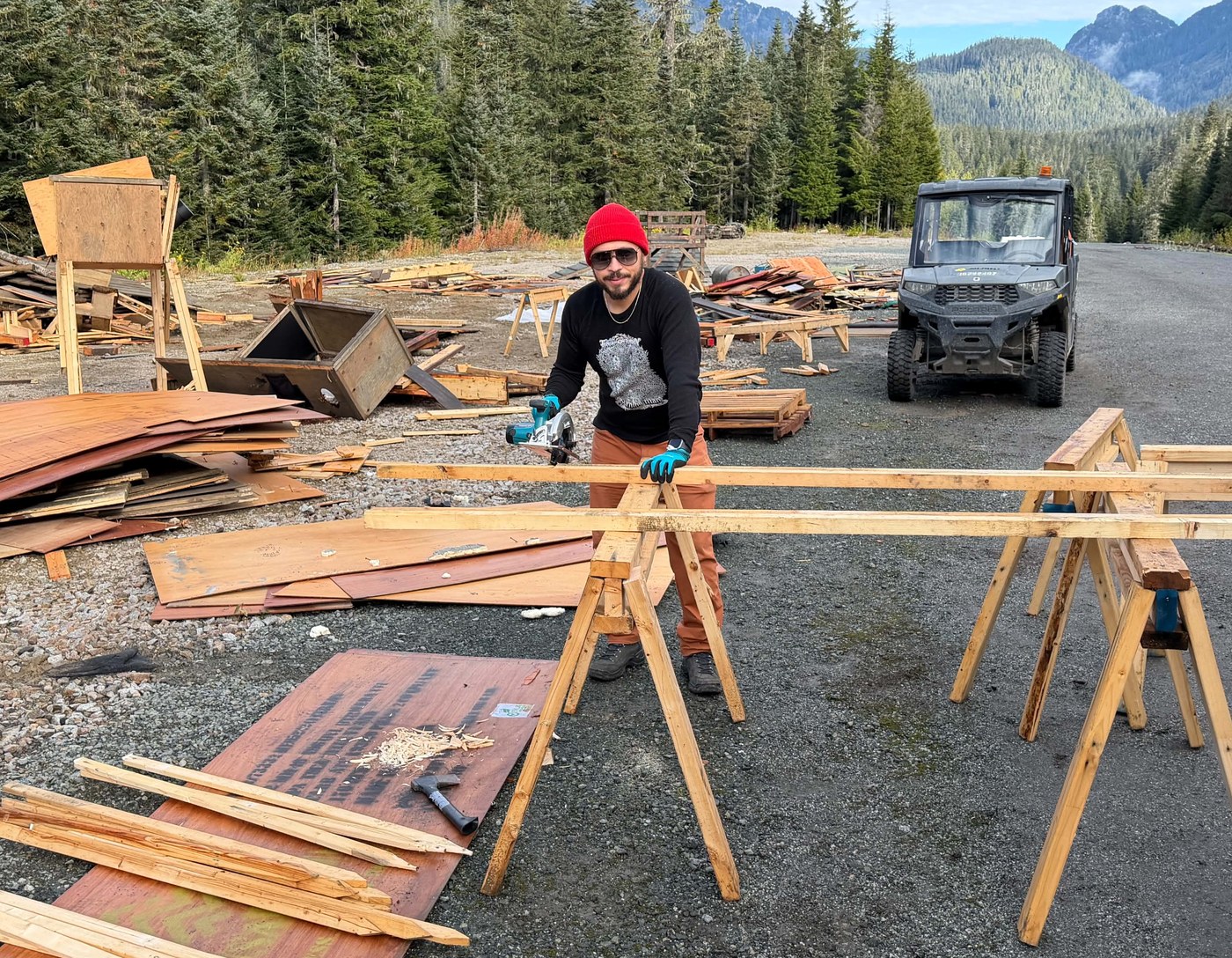 Ivan Otto in a red toque cutting lumber with a circular saw on sawhorses in a gravel lot, piles of plywood and a utility vehicle behind him